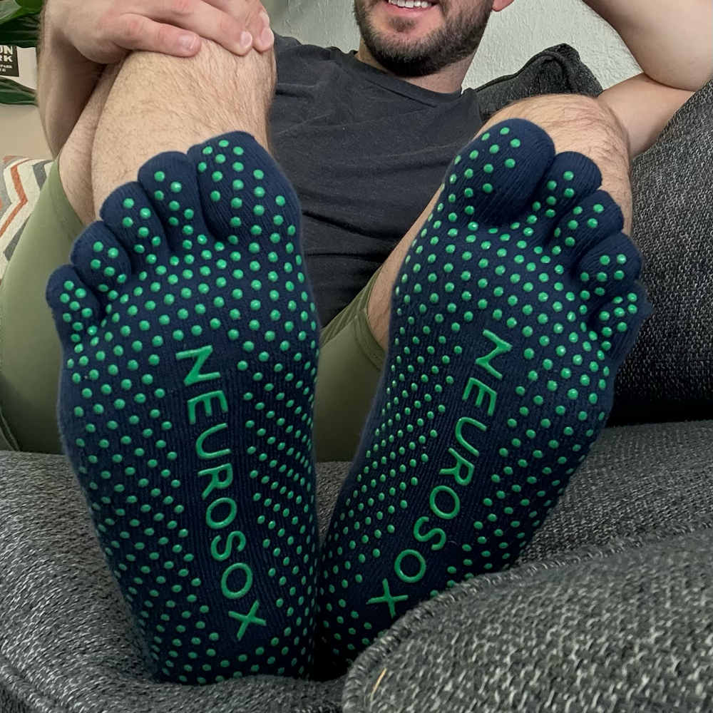 Man relaxing on couch wearing navy NeuroSox Five Toe Grip Socks, soles facing forward showing green anti-slip dots and logo