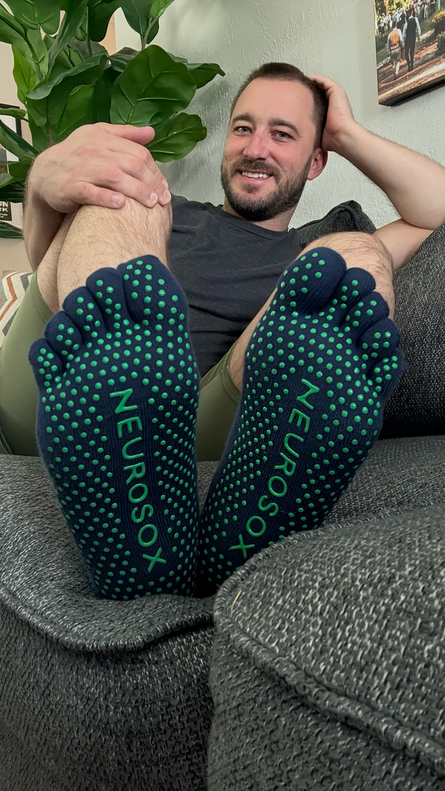 Man relaxing on couch wearing navy NeuroSox Five Toe Grip Socks, soles facing forward showing green anti-slip dots and logo