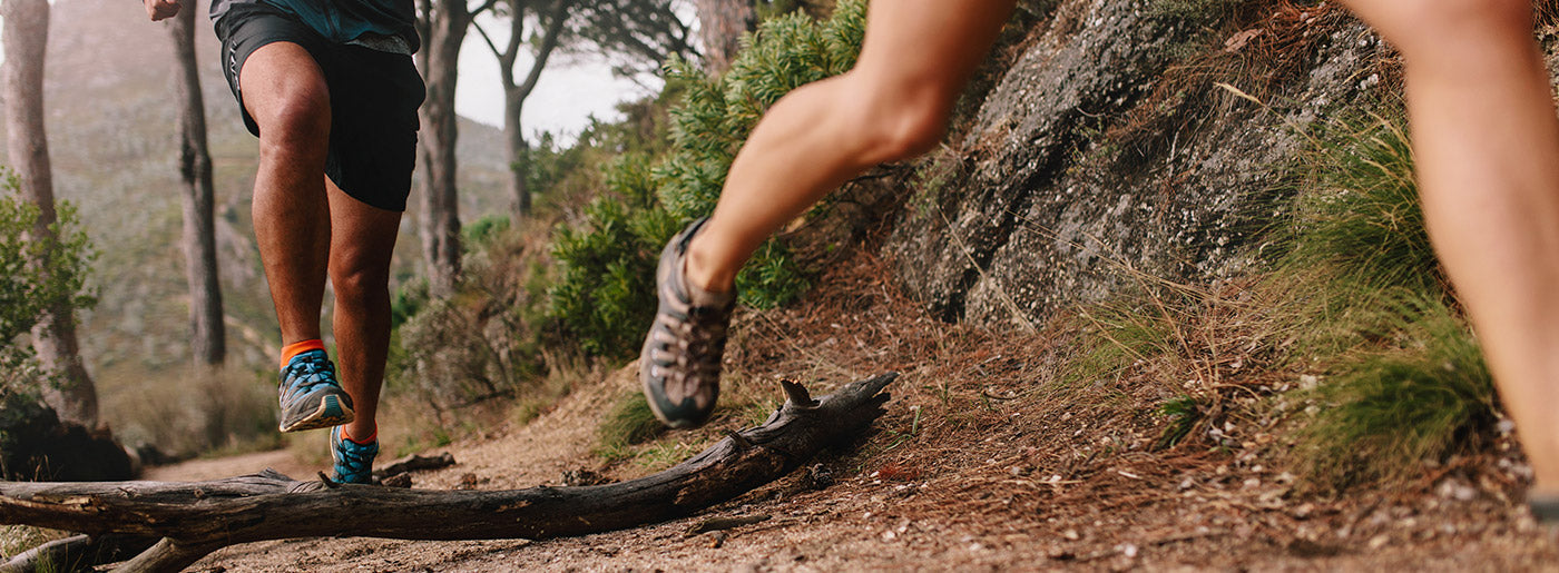 Trail runners jumping over log on mountain path, highlighting need for durable toe socks for hiking
