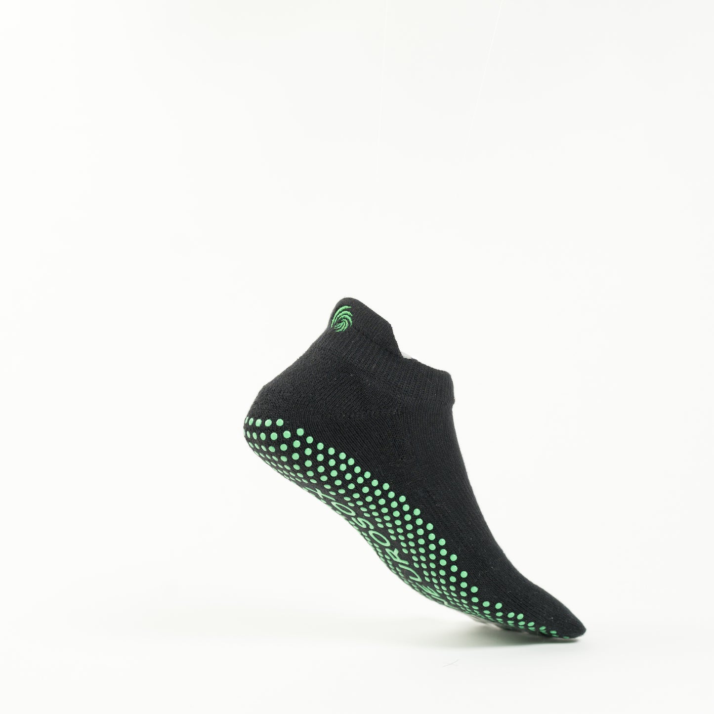 The image shows a single black colored sock with green grip dots on the sole, positioned as if suspended in mid-air against a plain white background. The sock has a small green swirl logo near the ankle area.