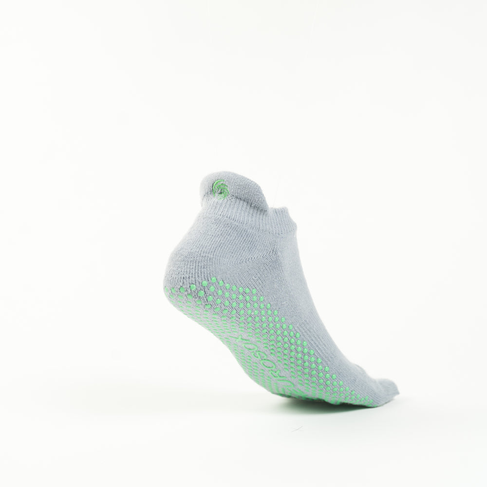 The image shows a single grey colored sock with green grip dots on the sole, positioned as if suspended in mid-air against a plain white background. The sock has a small green swirl logo near the ankle area.