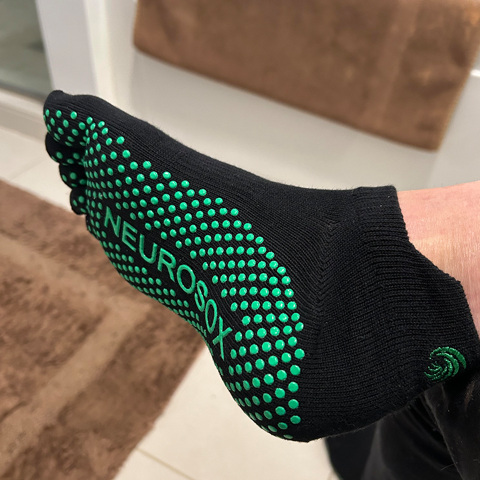 
                  
                    The image shows a close-up of a person’s foot wearing a black sock with green grip dots on the sole. The sock has the word "NEUROSOX" printed on the bottom in green. The background includes a bathroom setting, with a brown towel hanging over the edge of a white bathtub.
                  
                