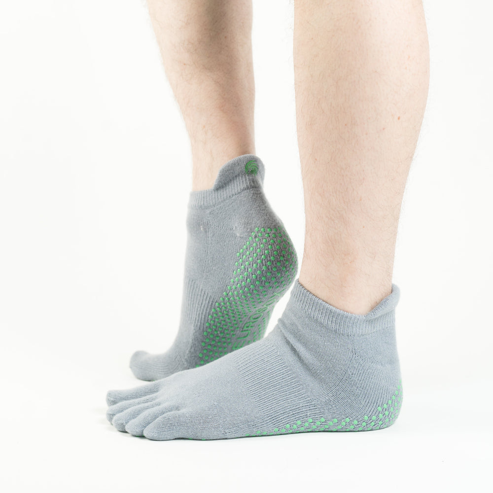 A pair of gray five-toe grip socks with green grip patterns on the sole, featuring a low-rise design with a raised heel tab, worn on a person's feet.