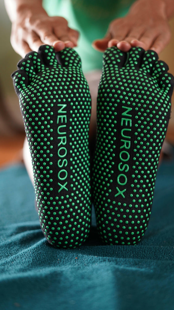 
                  
                    NeuroSox Five Toe Grip Socks with green anti-slip soles and logo visible during seated stretch on blue mat.
                  
                
