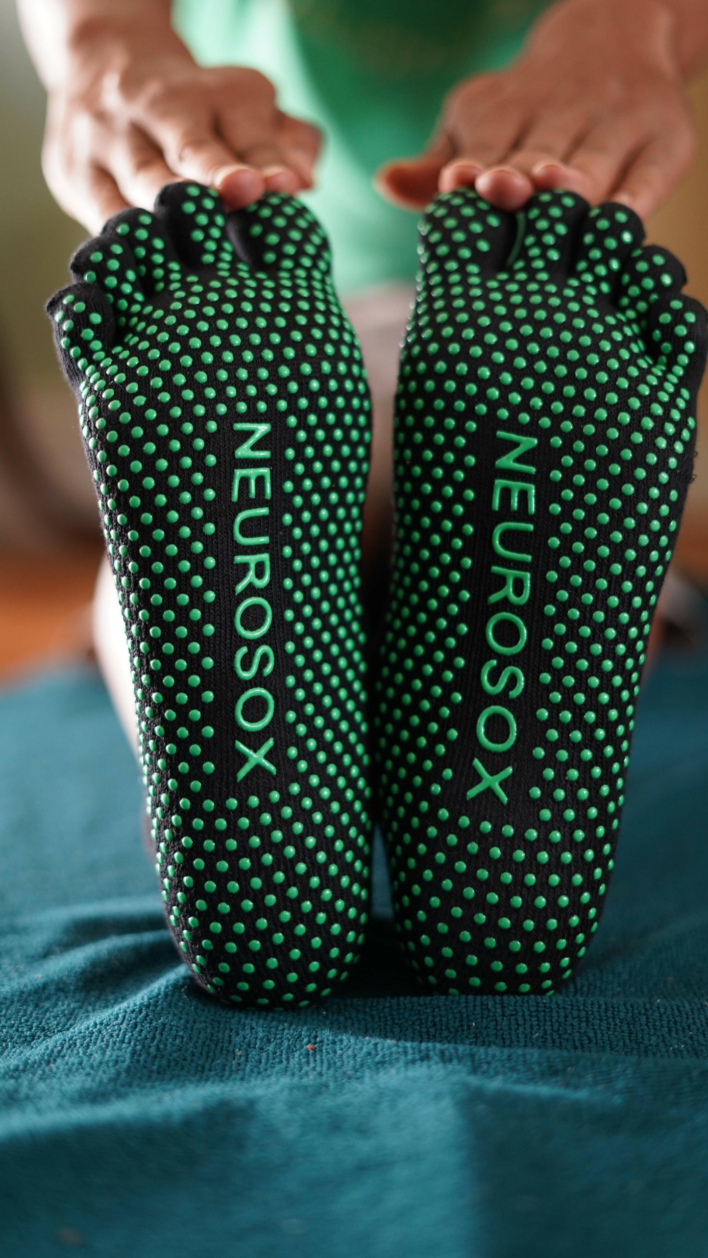 
                  
                    NeuroSox Five Toe Grip Socks with green anti-slip soles and logo visible during seated stretch on blue mat.
                  
                