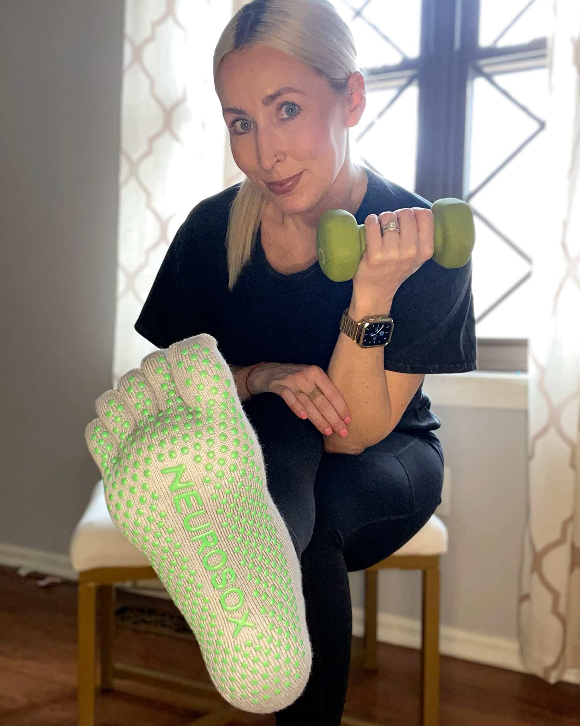 Woman sitting on chair holding a green dumbbell and wearing white NeuroSox with green grip dots, sole facing camera.